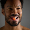 Man with a small orange object in his mouth against a gray background